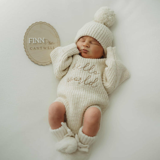Newborn baby in a knitted outfit and hat lying on a white surface with a BIRTH ANNOUNCEMENT PLAQUE sign.